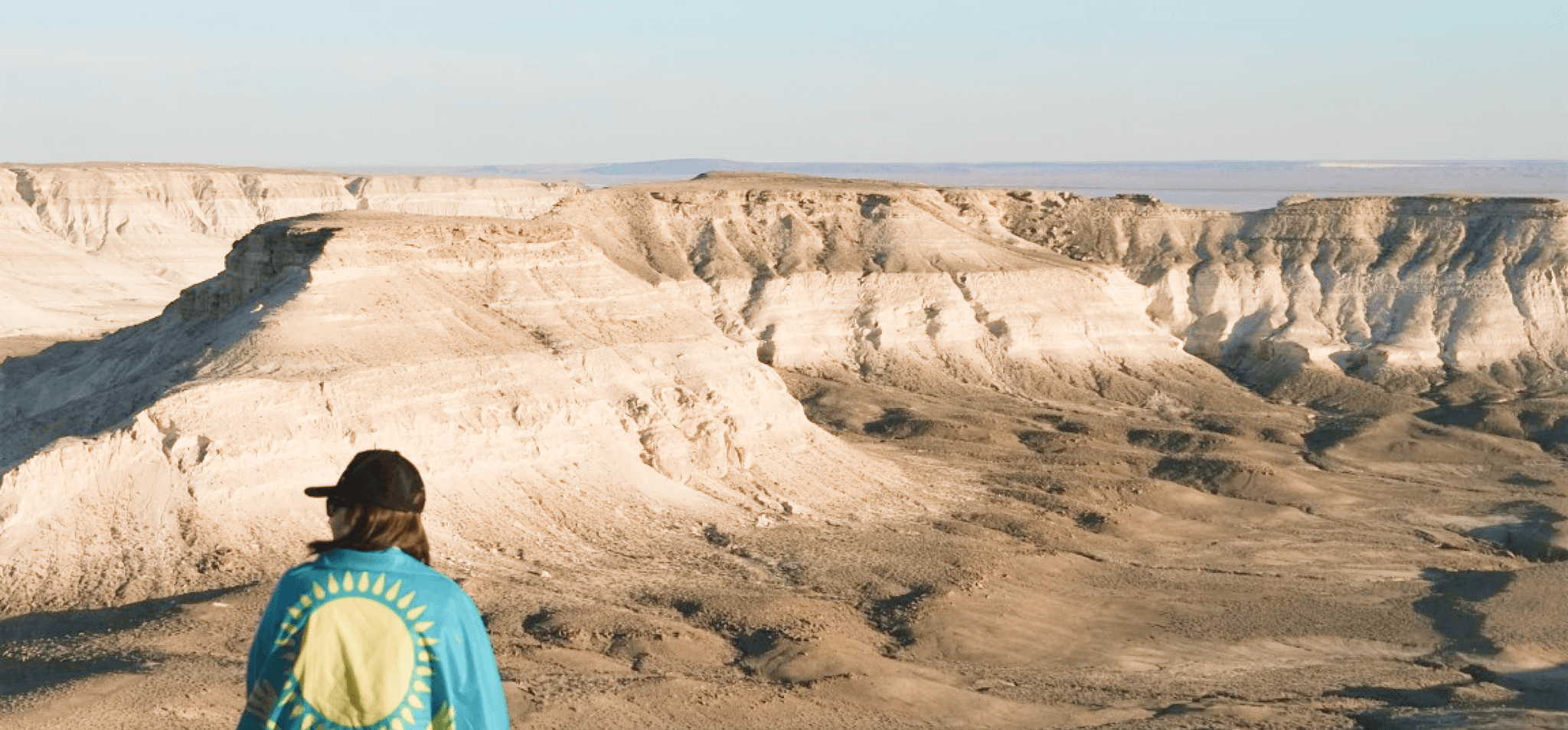 A photo of Charyn canyon in Kazakhstan and a girl gazing at it, with a flag of Kazakhstan on her back.